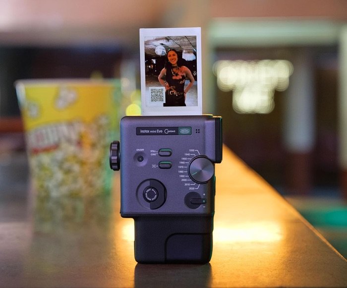 Cámara Fujifilm Instax Mini Evo imprimiendo una fotografía instantánea con código QR en un entorno de cine con palomitas al fondo.