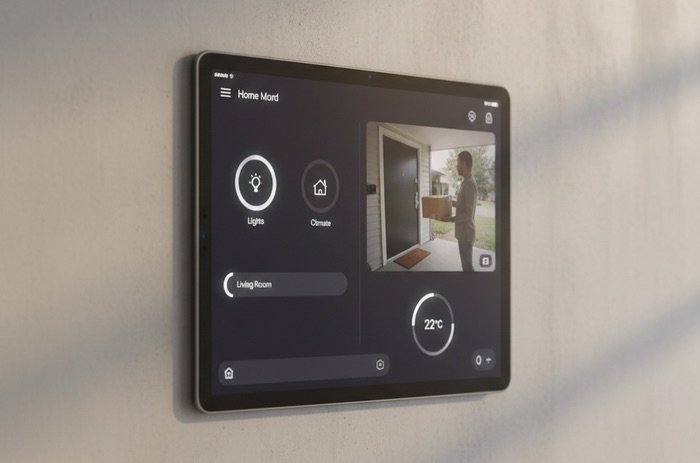 iPad montado en una pared minimalista mostrando una interfaz de hogar inteligente con control de luces, temperatura de 22 grados y vista de cámara de seguridad en tiempo real.